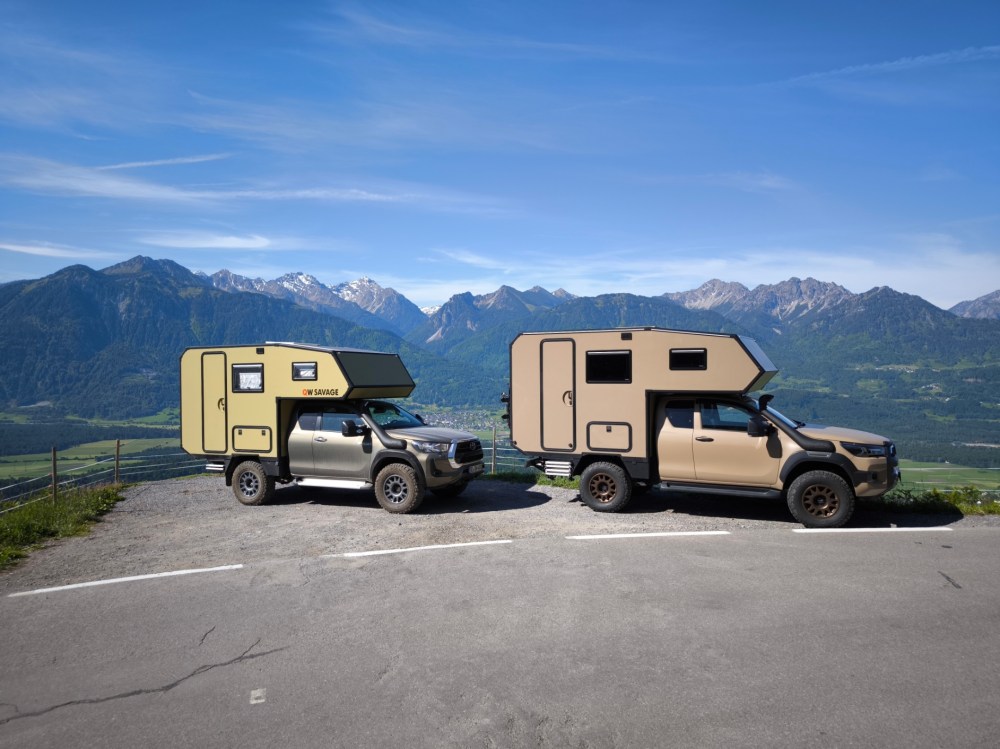 Zwei offroad-fähige Fahrzeuge mit Wohnkabinen auf einem Parkplatz mit Aussicht auf die Berge, unter einem blauen Himmel.