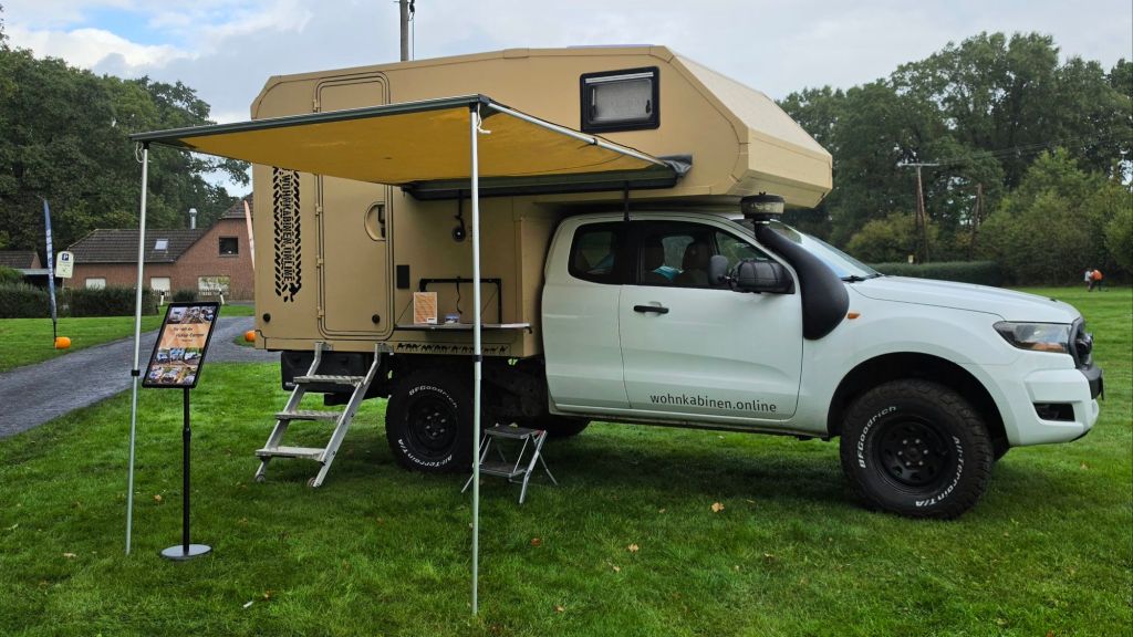 Ein Wohnmobil mit aufgesetztem Dach auf einem Pickup-Truck, unter einem ausklappbaren Sonnenschutz, auf einer Wiese. Neben dem Fahrzeug steht ein Informationsschild.
