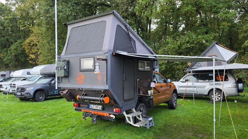 Ein Zeltanhänger mit einem erhöhten Dach, der auf einem Geländewagen steht, umgeben von anderen Fahrzeugen auf einer grünen Wiese.