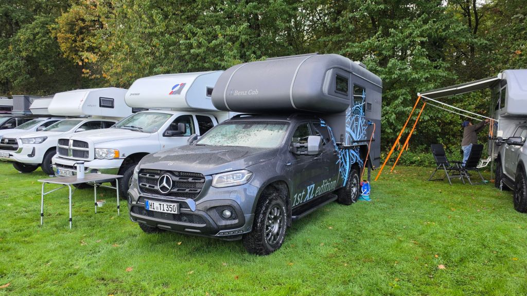 Ein Mercedes-Pickup mit Aufstelldach und Camper-Anhänger steht auf einer Wiese, umgeben von mehreren anderen Wohnmobilen und einem Tisch mit Stühlen.