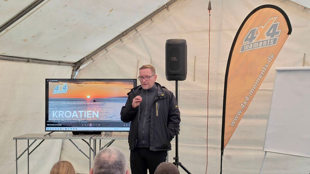 Ein Vortragender spricht vor einem Publikum in einem Zelt, während auf einem Bildschirm im Hintergrund eine Präsentation über Kroatien und Camper-Touren gezeigt wird.
