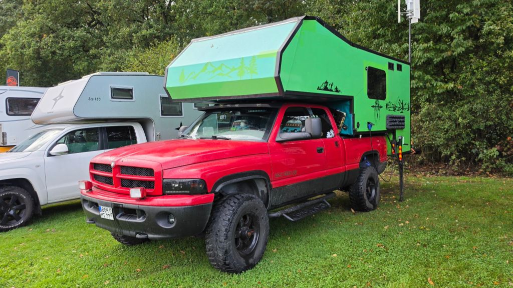 Ein roter Pickup-Truck mit einer grünen Aufsatzcamper. Der Camper hat eine moderne, rechteckige Form und ist mit Bergmotiven verziert. Im Hintergrund sind weitere Wohnmobile zu sehen.