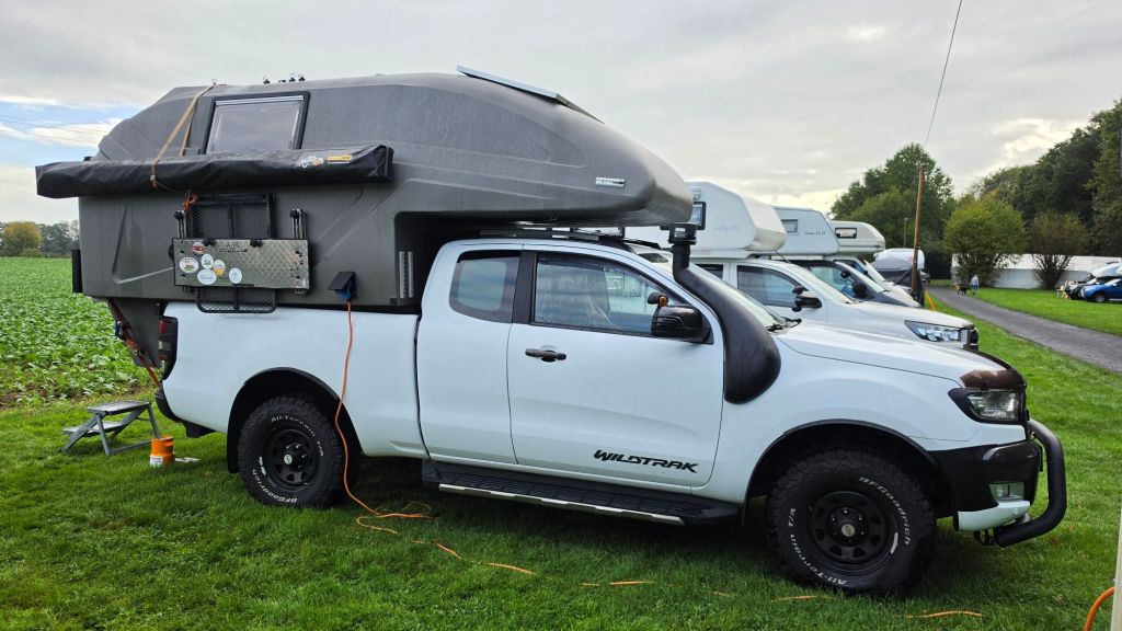 Ein weißer Pickup-Truck mit einem grauen Wohnmodul auf der Ladefläche, der auf einer Wiese steht, umgeben von anderen Wohnmobilen.