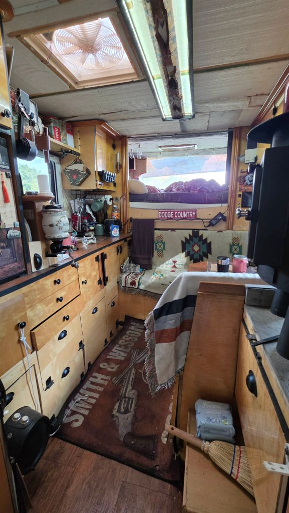 Blick ins Innere eines Wohnmobils mit Holzregalen, einer kleinen Küchenzeile und einem Bett im Hintergrund. Der Boden ist mit einem Teppich mit einem Pistolenmotiv ausgelegt, und es gibt eine Mischung aus modernen und rustikalen Dekorationen.