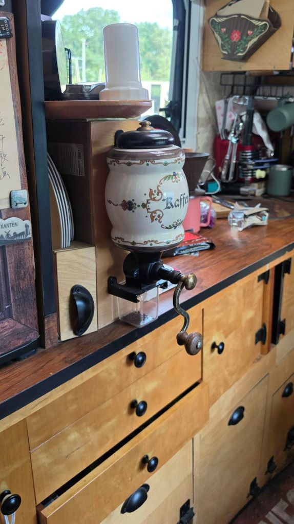 Ein antiker Kaffeebehälter mit der Aufschrift 'Kaffee' steht auf einem Holztisch in einer Küche. Der Behälter hat einen Handgriff und einen dekorativen Stil. Im Hintergrund sind Küchengeschirr und Utensilien sichtbar.