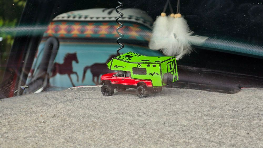 Ein Spielzeugauto in rot mit einem grünen Wohnmobil-Anhänger auf einer grauen Fläche im Innenraum eines Fahrzeugs.