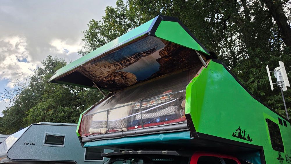 Ein grüner Campinganhänger mit einem geöffneten Dach, das eine Landschaftsaufzeichnung zeigt. Im Inneren sind gestapelte Bettlaken und Campingausrüstung sichtbar.