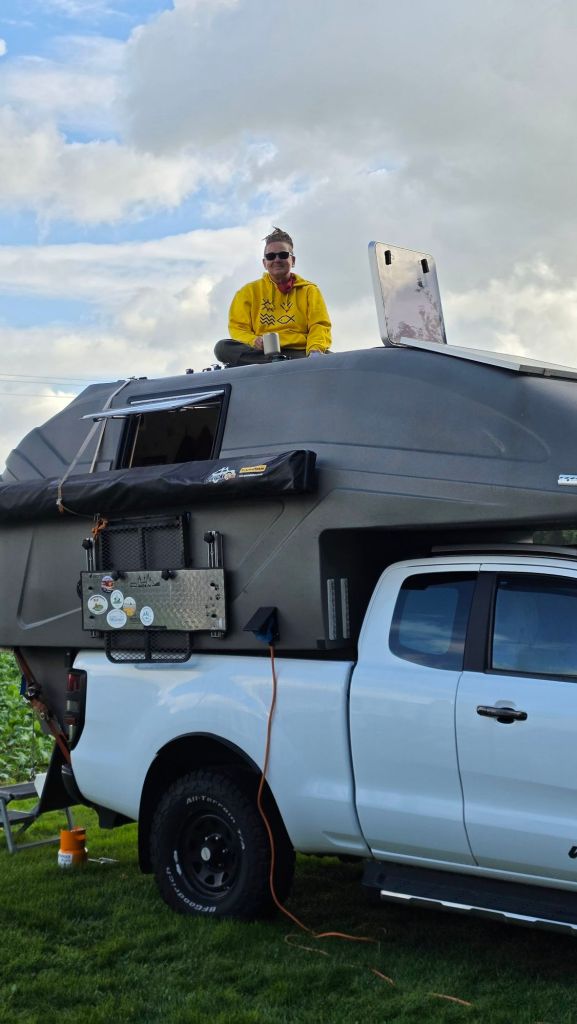 Eine Person sitzt auf dem Dach eines Wohnwagens, der auf einem weißen Pickup-Truck montiert ist. Im Hintergrund sind graue Wolken zu sehen.