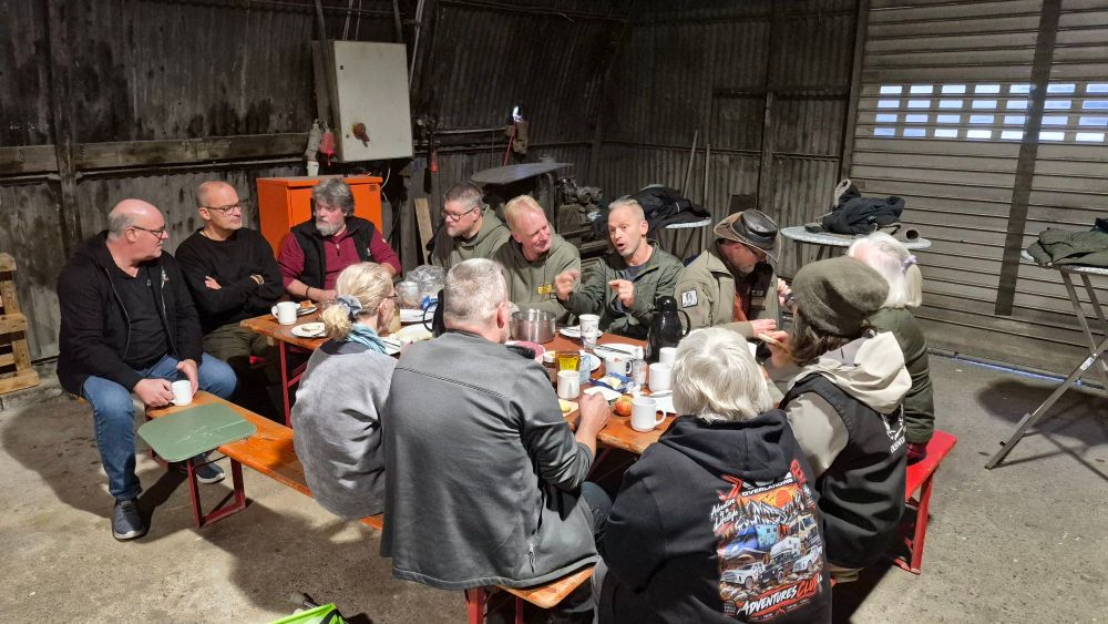 Eine Gruppe von Menschen sitzt an einem Tisch in einer Scheune und unterhält sich bei Snacks und Getränken.