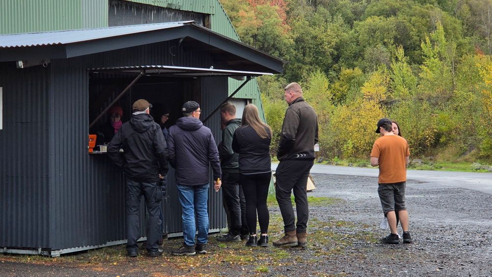 Eine Gruppe von Menschen steht an einem kleinen Verkaufsstand in der Natur, umgeben von Bäumen mit herbstlichem Laub.