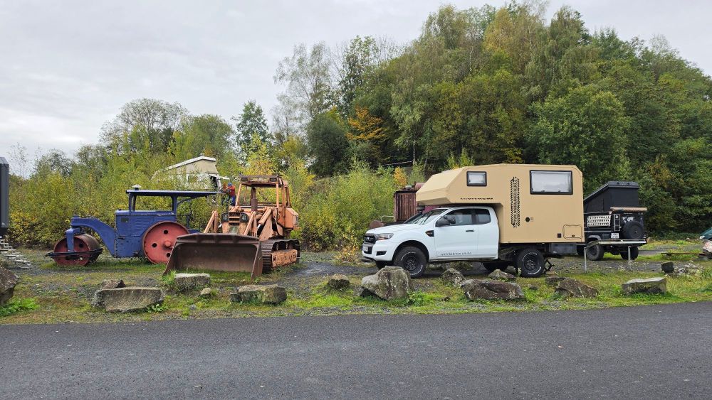 Eine Sammlung von alten landwirtschaftlichen Maschinen und einem modernen Wohnmobil auf einem Schotterplatz, umgeben von Bäumen.