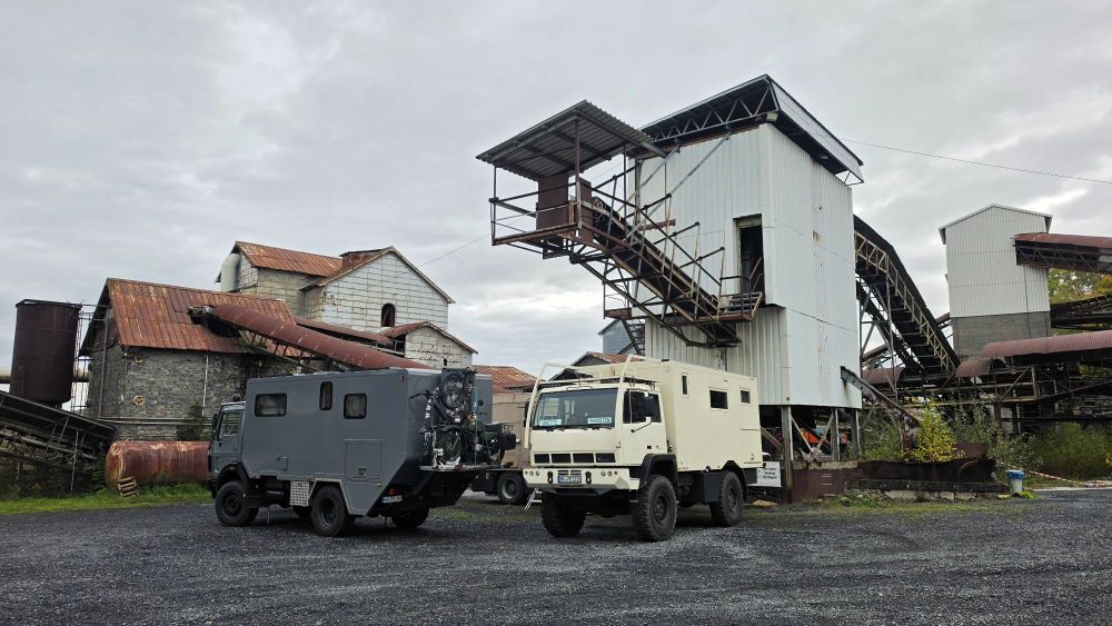 Zwei Militärfahrzeuge stehen auf einem Schotterplatz vor einer alten Industrieanlage mit mehreren Gebäuden und Förderbändern.