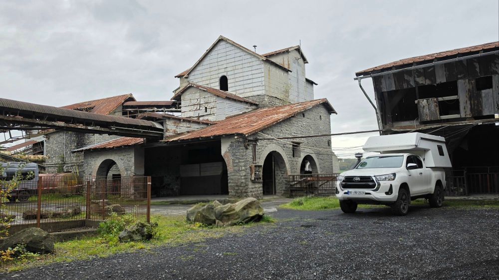 Eine weiße Fahrzeug mit Wohnkabine steht vor einer alten, teilweise abgerissenen Fabrikgebäudekomplex mit einer Mischung aus Stein- und Metallstrukturen. Im Hintergrund sind abgenutzte Dächer und Förderbänder zu sehen. Der Himmel ist bewölkt.