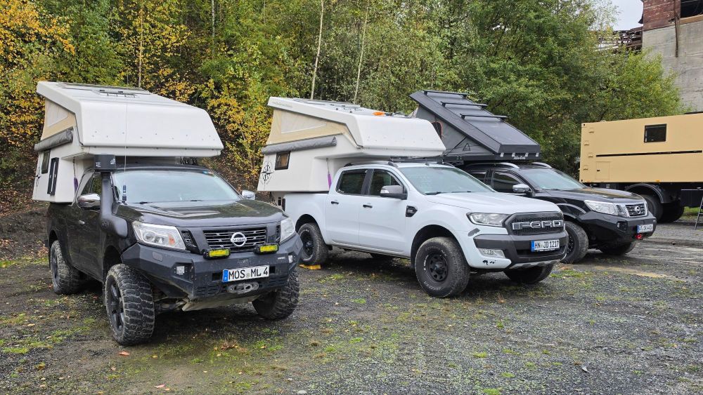 Drei Offroad-Fahrzeuge mit Campingaufbauten parken in einer ländlichen Umgebung mit Bäumen im Hintergrund.