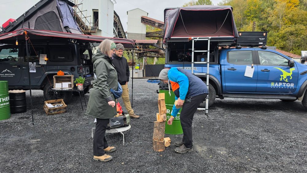 Drei Personen stehen an einem Campingplatz mit Wohnmobilen. Eine Frau in einem grünen Mantel spricht mit einem Mann, während ein anderer Mann mit einem blauen Oberteil Holz spaltet.
