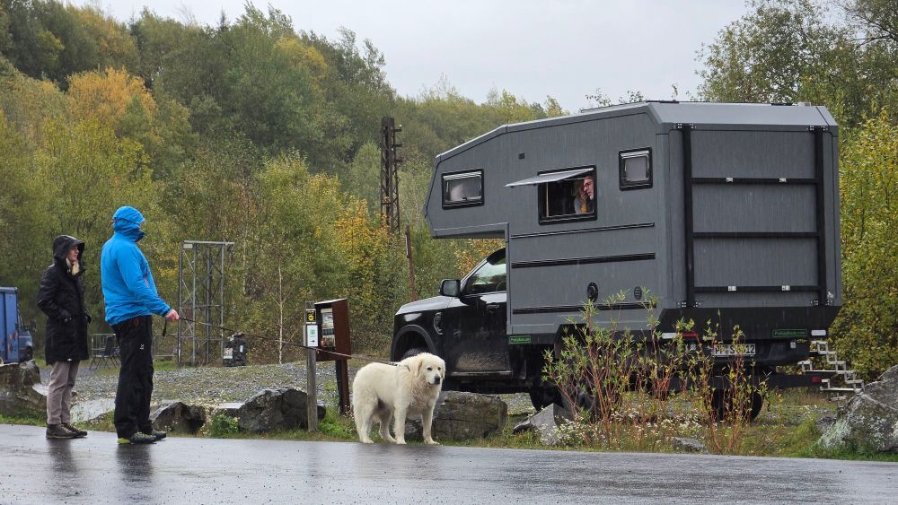 Zwei Personen stehen bei einem Wohnmobil, während ein Hund daneben sitzt. Im Hintergrund sind Bäume und eine regnerische Landschaft zu sehen.