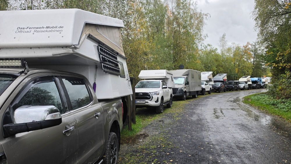 Reihe von Wohnmobilen und Offroad-Reisekabinen auf einem schmalen Weg in einer bewaldeten Umgebung, leicht regnerisches Wetter.