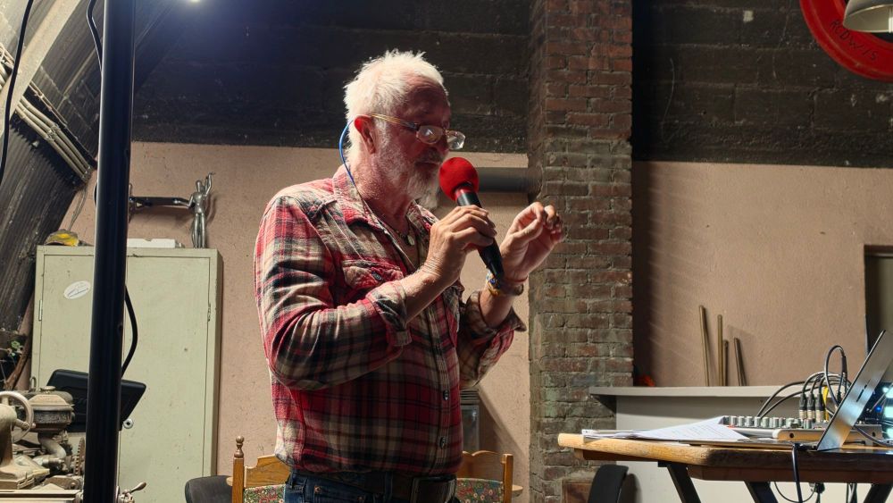 Ein älterer Mann mit Brille und einem karierten Hemd spricht mit einem Mikrofon in der Hand. Im Hintergrund sind ein Tisch, technische Geräte und eine Wand mit Mauerwerk zu sehen.