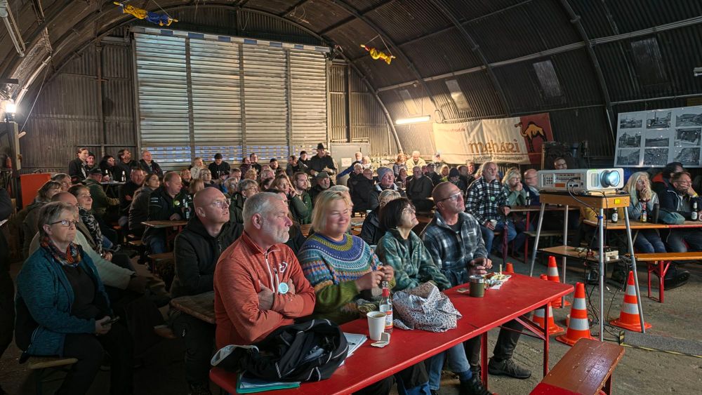 Eine große Gruppe von Menschen sitzt in einem Innenraum mit rotem Tisch und projektiertem Bild, während sie aufmerksam lauschen.
