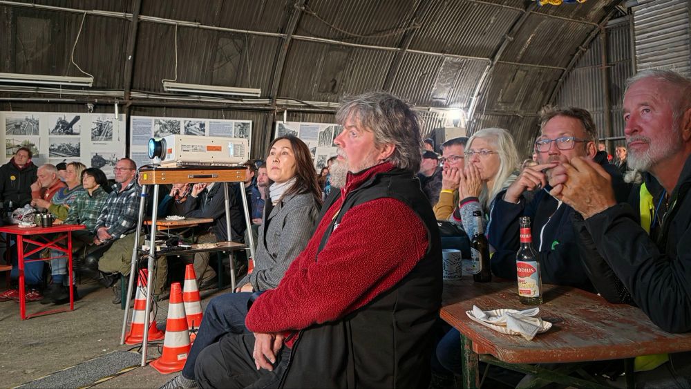 Ein Publikum sitzt in einem Raum mit einer Leinwand, während ein Projektor in der Mitte der Ansicht steht. Die Zuschauer blicken interessiert und aufmerksam auf das Geschehen vor ihnen. Verschiedene Getränke und Snacks sind auf den Tischen vorhanden.
