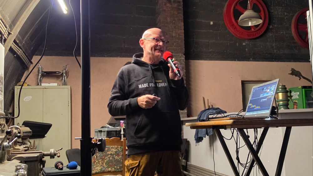 Ein Mann mit Brille und Mikrofon spricht auf einer Veranstaltung, während er auf einen Laptop zeigt. Im Hintergrund sind verschiedene Requisiten und eine Lampe sichtbar.