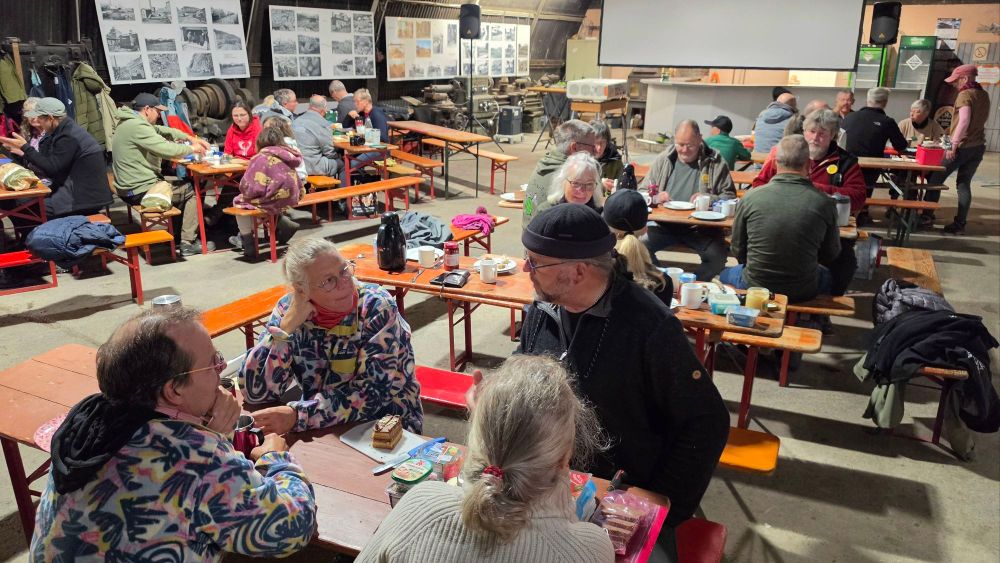 Eine Gruppenveranstaltung in einer Halle, bei der Menschen an langen Tischen sitzen, essen und sich unterhalten. Im Hintergrund sind historische Fotos displayed.