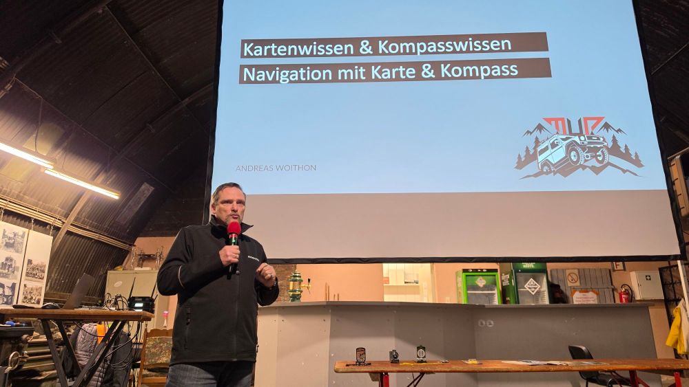 Ein Mann steht auf der Bühne und spricht mit einem Mikrofon, während eine Präsentation über Karten- und Kompasswissen im Hintergrund angezeigt wird.
