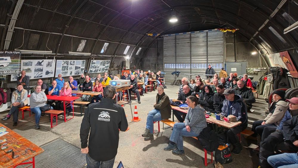 Eine Gruppe von Menschen hört einem Vortrag in einer großen Halle zu, die mit Tischen und Stühlen ausgestattet ist. An den Wänden hängen zahlreiche Bilder. Ein Referent steht vorne und präsentiert auf einem Bildschirm.