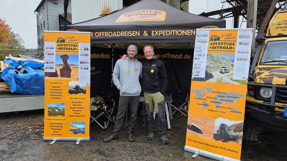Zwei Männer stehen vor einem Stand mit dem Banner 'ADVENTURE OFFROAD', der Informationen über Offroad-Reisen und Expeditionen zeigt. Im Hintergrund sind Fahrzeuge und Materialien sichtbar.