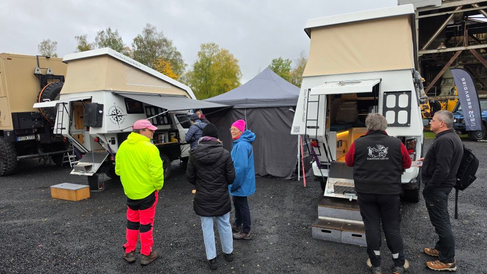 Gruppe von Menschen, die sich um Wohnmobile und ein Zelt bei einer Veranstaltung versammeln. Einige Personen tragen leuchtende Kleidung und diskutieren über die Fahrzeuge.