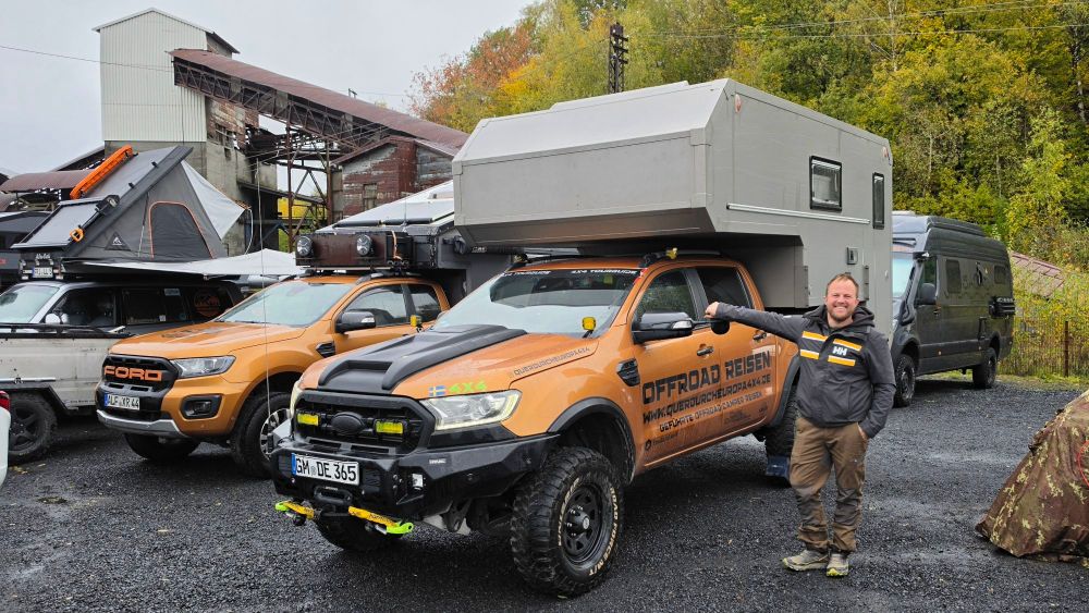 Ein Mann steht neben einem orangefarbenen Offroad-Fahrzeug mit einem Wohnaufbau, umgeben von weiteren Fahrzeugen und einer Industrieanlage im Hintergrund.