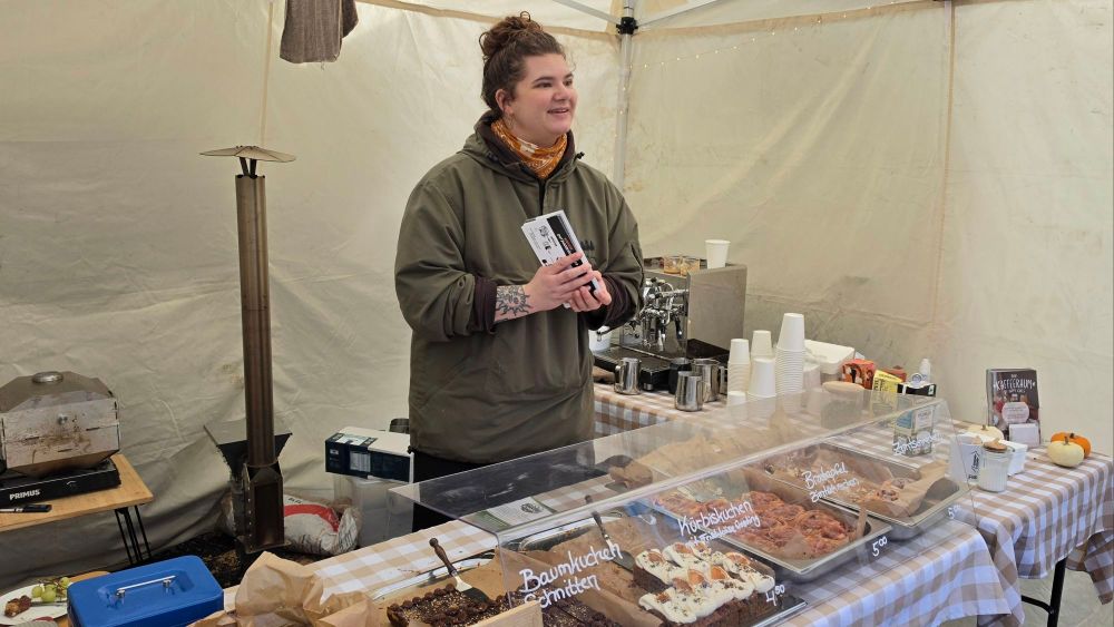 Eine Verkäuferin steht an einem Stand innerhalb eines Zeltes und hält ein Kassenbuch. Vor ihr ist eine Auswahl an Kuchen und Gebäcken in einer Vitrine aus Glas. Im Hintergrund sind Kaffeeausrüstung und Zubehör zu sehen.