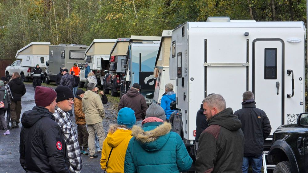 Menschen stehen vor einer Reihe von Wohnmobilen auf einem Parkplatz in der Natur, umgeben von Bäumen und Herbstblättern.