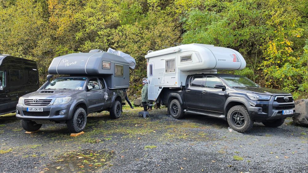 Zwei Wohnmobile stehen geparkt auf einem Kiesplatz umgeben von Bäumen. Das linke Fahrzeug hat eine graue Wohnkabine, das rechte Fahrzeug ist ein schwarzer Pick-up mit einem Campingaufbau.