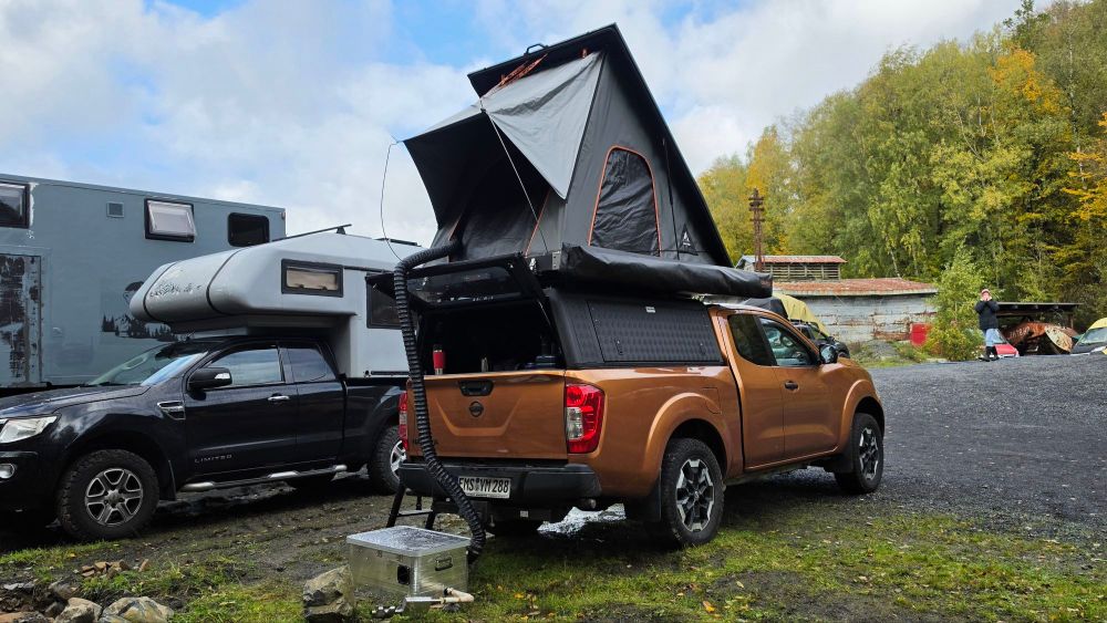 Ein orangefarbenes Pickup-Fahrzeug mit einem aufgeklappten Zelt auf der Ladefläche, geparkt auf einem unebenen Gelände neben einem schwarzen SUV und einem Wohnmobil im Hintergrund.