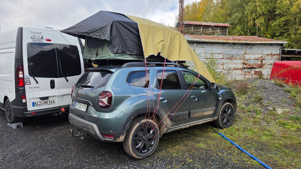 Ein grünes SUV mit einem ausgeklappten Dachzelt, das an einem weißen Lieferwagen geparkt ist. Im Hintergrund sind verrostete Strukturen und Bäume zu sehen.