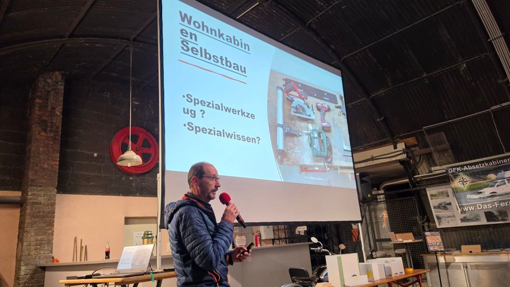 Ein Vortrag über den Selbstbau von Wohnkabinen, mit einem Referenten, der ein Mikrofon hält. Im Hintergrund eine Präsentationsfolie mit dem Titel "Wohnkabinen Selbstbau" und Fragen zu Spezialwerkzeugen und spezialisiertem Wissen. Auf der Folie sind Bilder von Werkzeugen zu sehen.