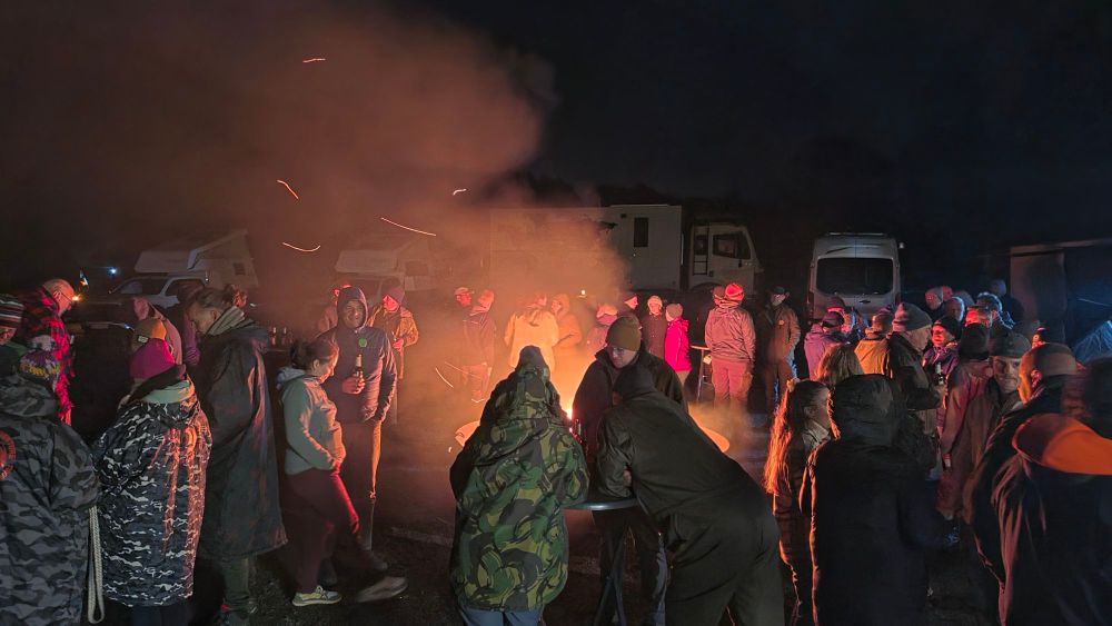 Eine Gruppe von Menschen versammelt sich um ein Feuer in der Nacht, Rauch und Funken steigen auf, während sie sich warm anziehen.
