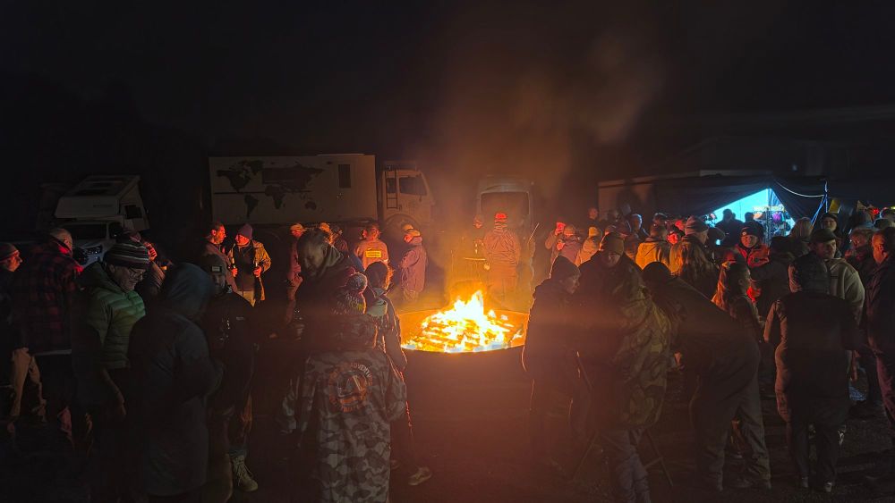 Menschen versammeln sich um ein großes Lagerfeuer in der Nacht, während Wohnmobile im Hintergrund stehen.
