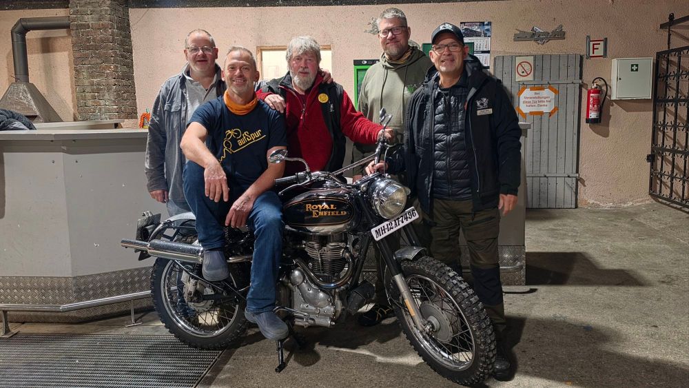 Fünf Männer stehen zusammen und posieren bei Nacht, während einer von ihnen auf einer Royal Enfield-Motorrad sitzt.