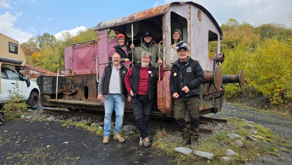 Gruppe von fünf Personen steht vor einer alten Lokomotive in der Natur, umgeben von Bäumen und Himmel.