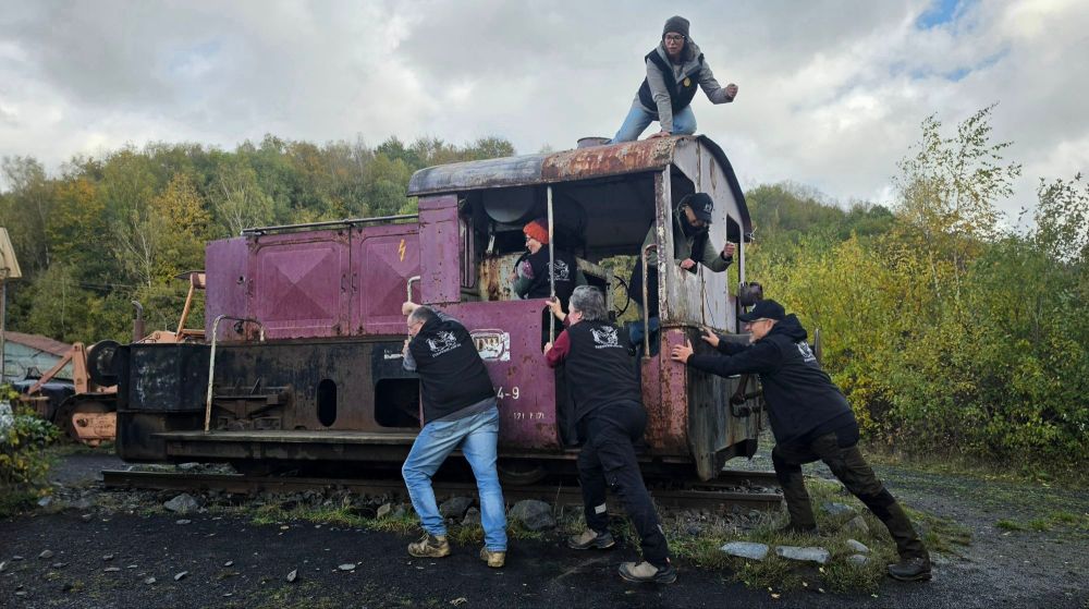 Eine Gruppe von fünf Personen, die an einer rostigen Lokomotive in der Natur herumklettern. Eine Person steht auf dem Dach der Lokomotive, während andere versuchen, in die Fenster zu klettern.