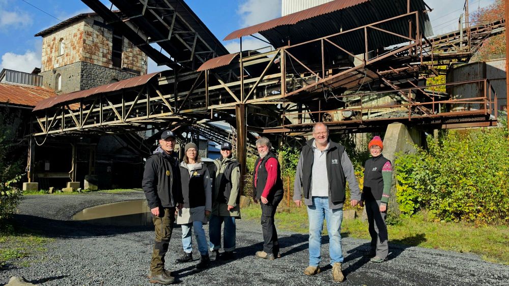 Sechs Personen stehen vor einer alten industriellen Anlage mit einem Förderband im Hintergrund. Die Gruppe ist sonnig und klar gekleidet, umgeben von grüner Vegetation.