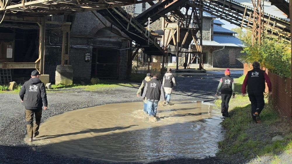 Gruppe von Personen läuft durch eine Pfütze auf einem unbefestigten Weg, im Hintergrund ist eine industrielle Struktur zu sehen.