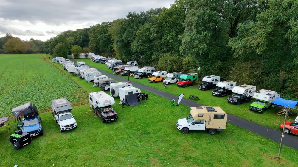 Luftaufnahme eines Campingplatzes mit mehreren Wohnmobilen, die auf einer grünen Wiese stehen, umgeben von Bäumen. Einige Fahrzeuge haben Markisen und Tische im Freien.