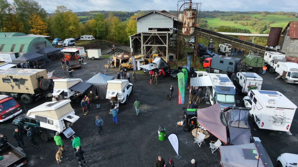 Luftaufnahme eines Outdoor-Messegeländes mit verschiedenen Wohnmobilen und Zelten, umgeben von Bäumen und ehemaliger industrieller Infrastruktur.