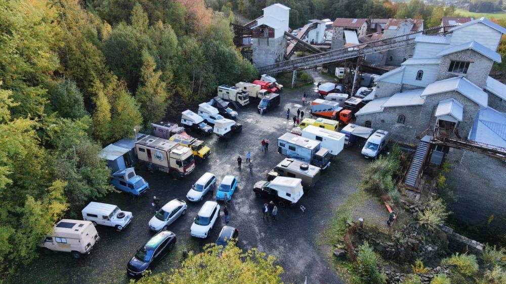 Luftaufnahme eines Parkplatzes mit verschiedenen Wohnmobilen und Autos, umgeben von Bäumen und industriellen Gebäuden.