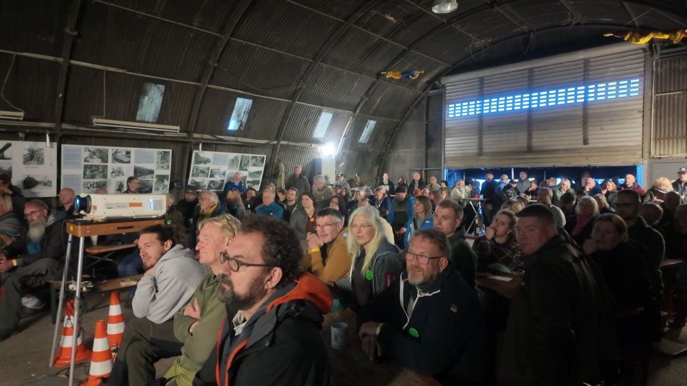 Eine große Gruppe von Menschen sitzt in einer Halle und schaut auf eine Präsentation, während einige Informationen an den Wänden präsentiert werden.