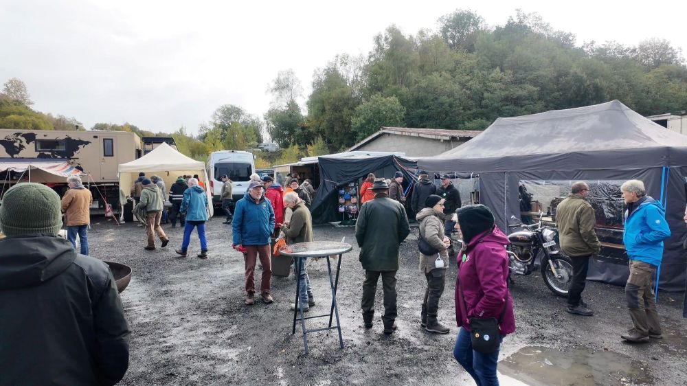 Eine belebte Outdoor-Veranstaltung mit mehreren Ständen und Zelten, wo Menschen in wetterfester Kleidung interagieren. Im Hintergrund sind Wohnmobile und Bäume zu sehen.