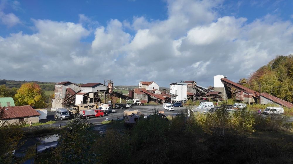 Luftaufnahme einer alten Industrieanlage mit mehreren Gebäuden und Förderbändern, umgeben von Fahrzeugen und Bäumen unter einem bewölkten Himmel.